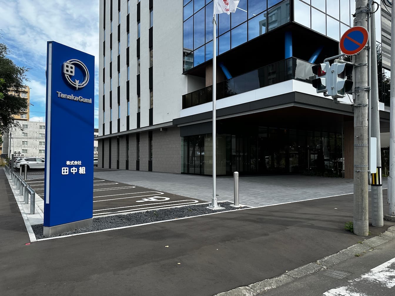 会社名の看板が立つ本社の外観。手前に来客用駐車場、奥のひさしの下にガラス張りの正面玄関がある。